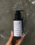 A hand holds a black pump bottle of Ives & Bees Facial Cream Wash against a wet, reflective gray concrete background.