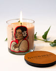 A lit candle in a clear glass jar with a beige wax body and a colorful illustrated label, shown beside a round wooden lid engraved with 'IVES & BEES'. Green leaves are placed around the candle on a clean white background.
