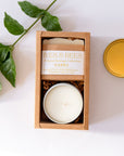 A boxed candle set on a white background with a green leaf sprig and a gold tin lid nearby. The main product is a rectangular kraft paper box containing a round tin candle and a wrapped soap bar.