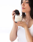 A woman holds a glass jar candle with cream-colored wax and a black lid, with the brand name 'IVES & BEES' printed on the glass, against a solid white background.
