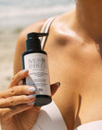 A person outdoors at the beach holds a black pump bottle labeled Ives & Bees Mineral & Plant Rose Sunscreen.