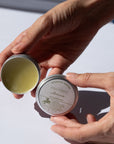Two hands holding Arnica Salve  against a light background.