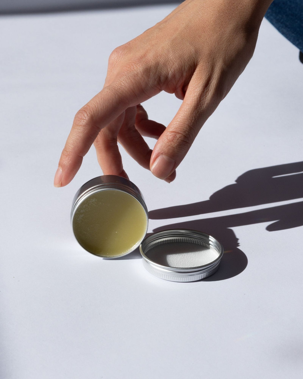 Hand holding a Arnica Salve with a lid, filled with a greenish substance on a light background.