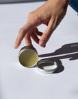 Hand holding a Arnica Salve with a lid, filled with a greenish substance on a light background.