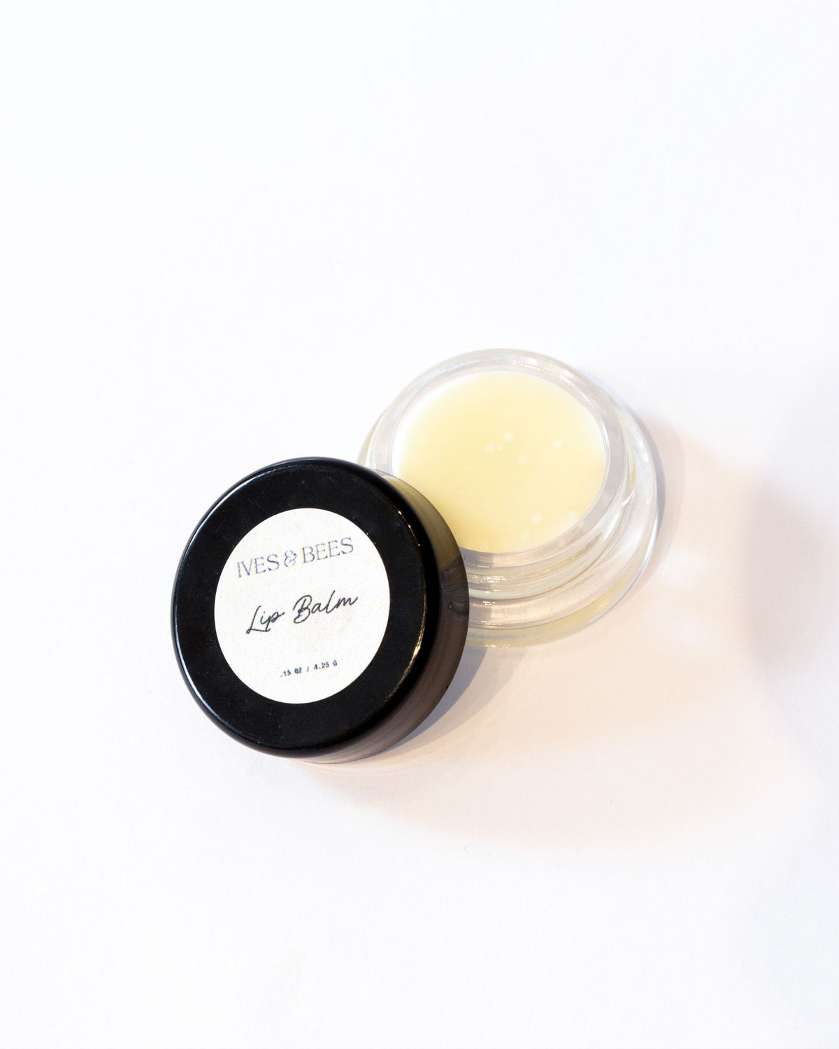 A small open jar of lip balm with pale yellow balm visible inside, next to its black lid which has a white label reading 'Ives & Bees Lip Balm'.