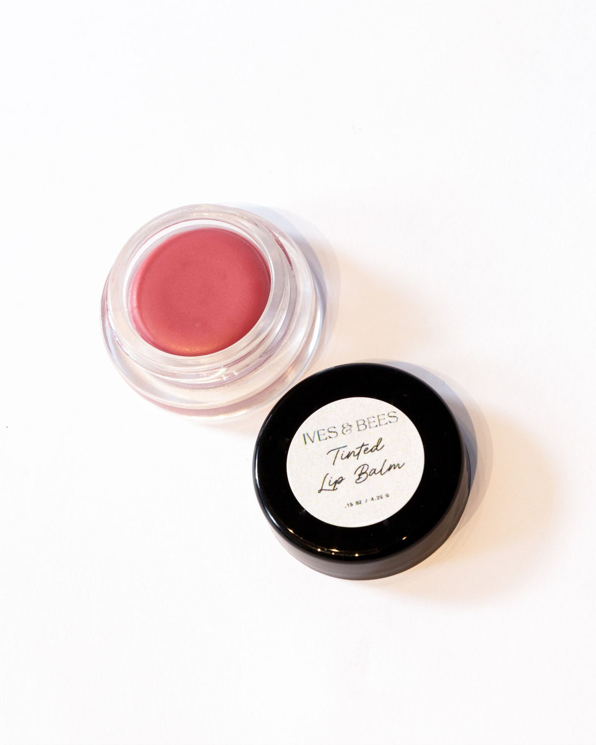 Ives & bees Tinted Lip Balm in a clear round pot showing the pinkish-red balm, with its black lid featuring a white label placed beside it on a clean white background.