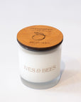 A white candle in a clear glass jar with a round wooden lid engraved with a lemon illustration and the brand name 'IVES & BEES'.