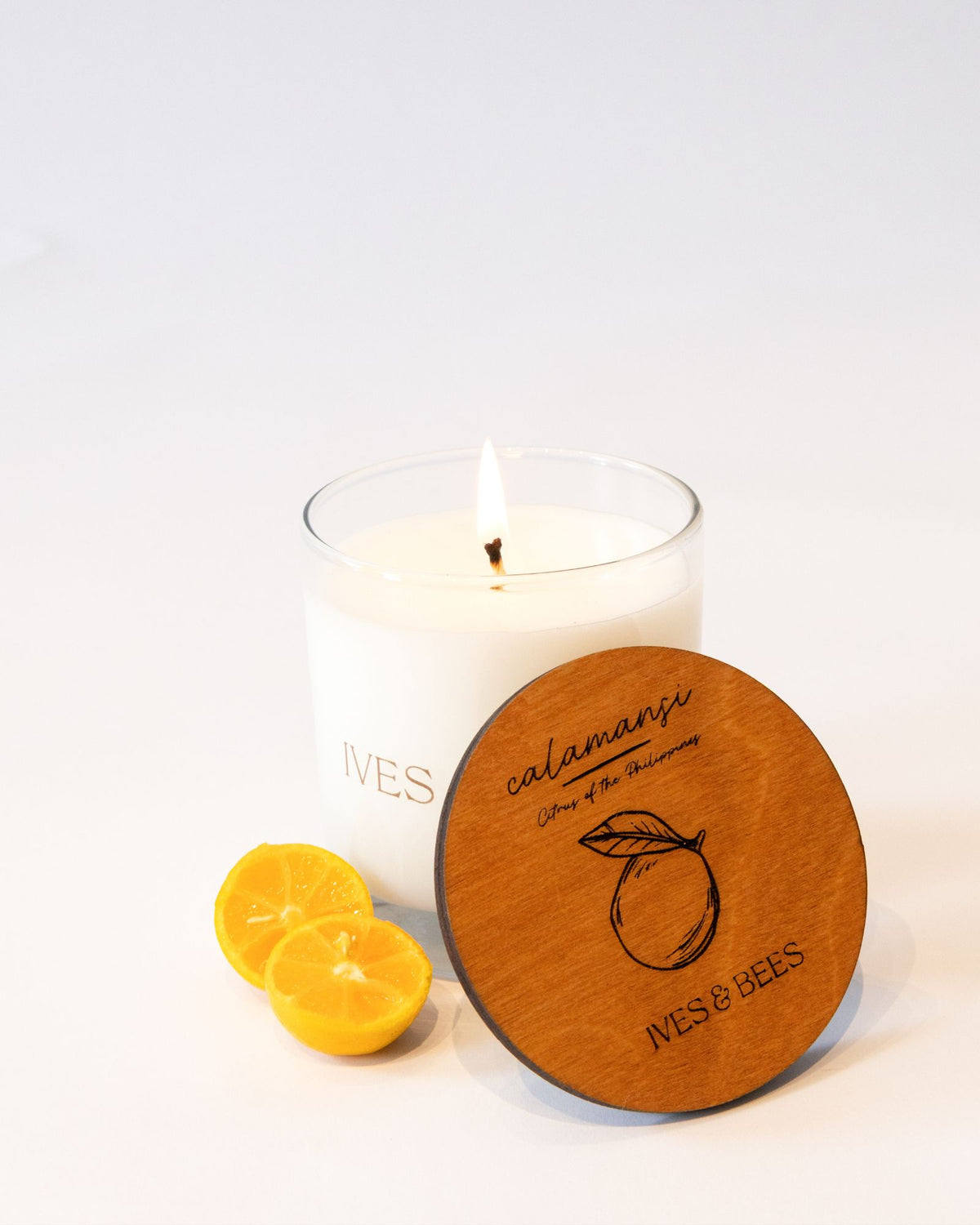 A white candle in a clear glass jar with a lit flame, shown on a white background with two lemon slices and a round wooden lid in front. The lid is engraved with 'calamansi', 'Scent of the Seasoning', and 'IVES & BEES'.