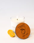 A white candle in a clear glass jar with a lit flame, shown on a white background with two lemon slices and a round wooden lid in front. The lid is engraved with 'calamansi', 'Scent of the Seasoning', and 'IVES & BEES'.