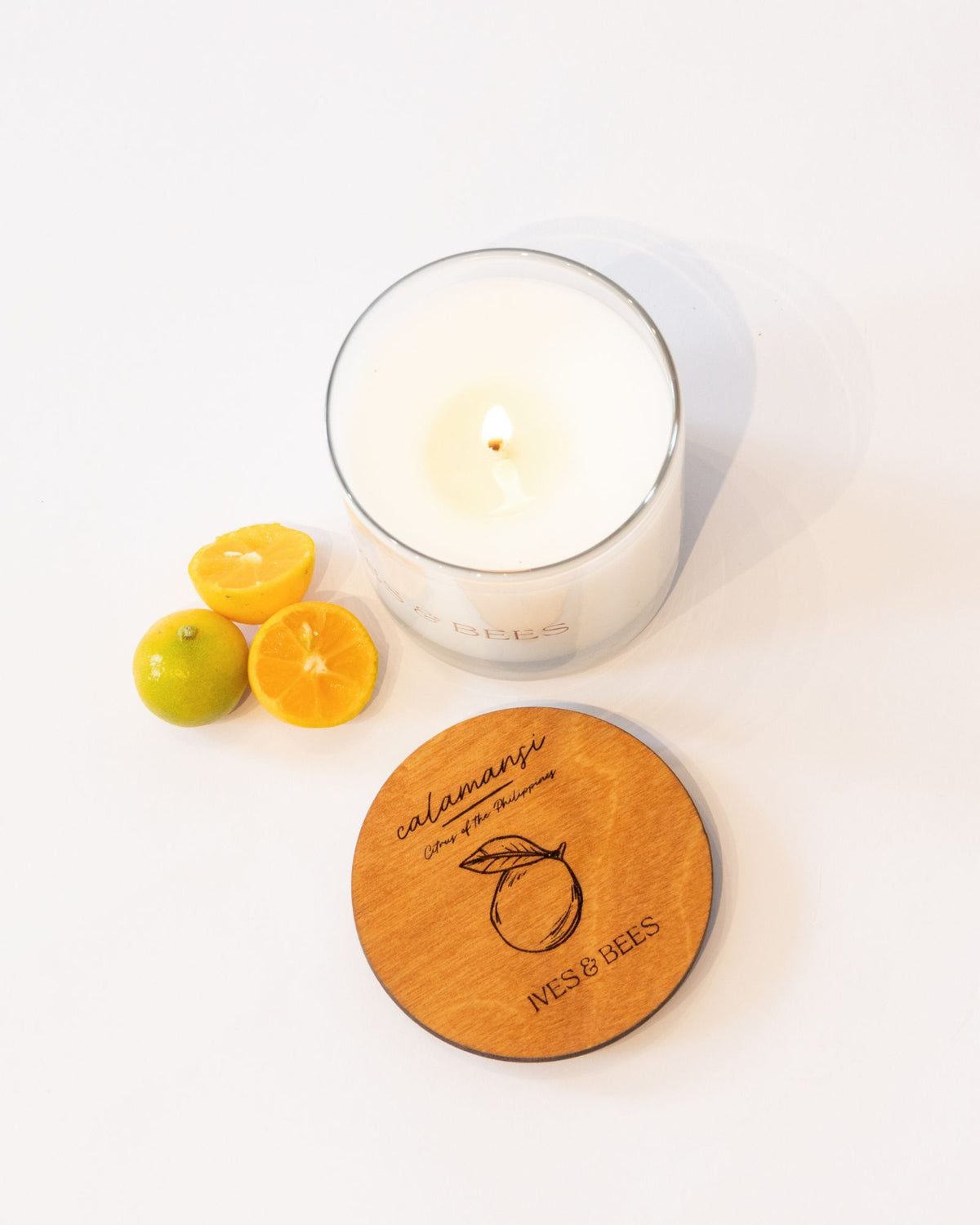 A lit white candle in a clear glass jar sits on a white surface beside a round wooden lid engraved with the scent name 'calamansi' and 'WEST OF EES,' plus three small citrus fruits.