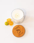 A lit white candle in a clear glass jar sits on a white surface beside a round wooden lid engraved with the scent name 'calamansi' and 'WEST OF EES,' plus three small citrus fruits.