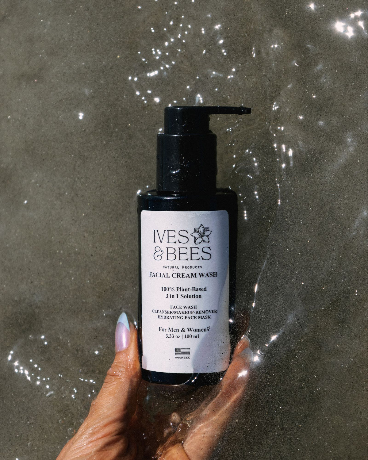 A hand holds a black pump bottle of Ives & Bees Facial Cream Wash against a wet, reflective gray concrete background.