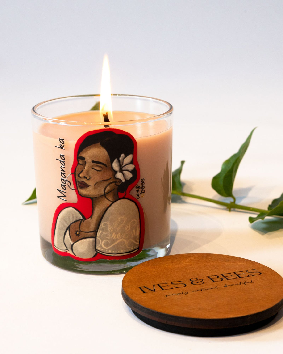 A lit candle in a clear glass jar with a beige wax body and a colorful illustrated label, shown beside a round wooden lid engraved with 'IVES & BEES'. Green leaves are placed around the candle on a clean white background.