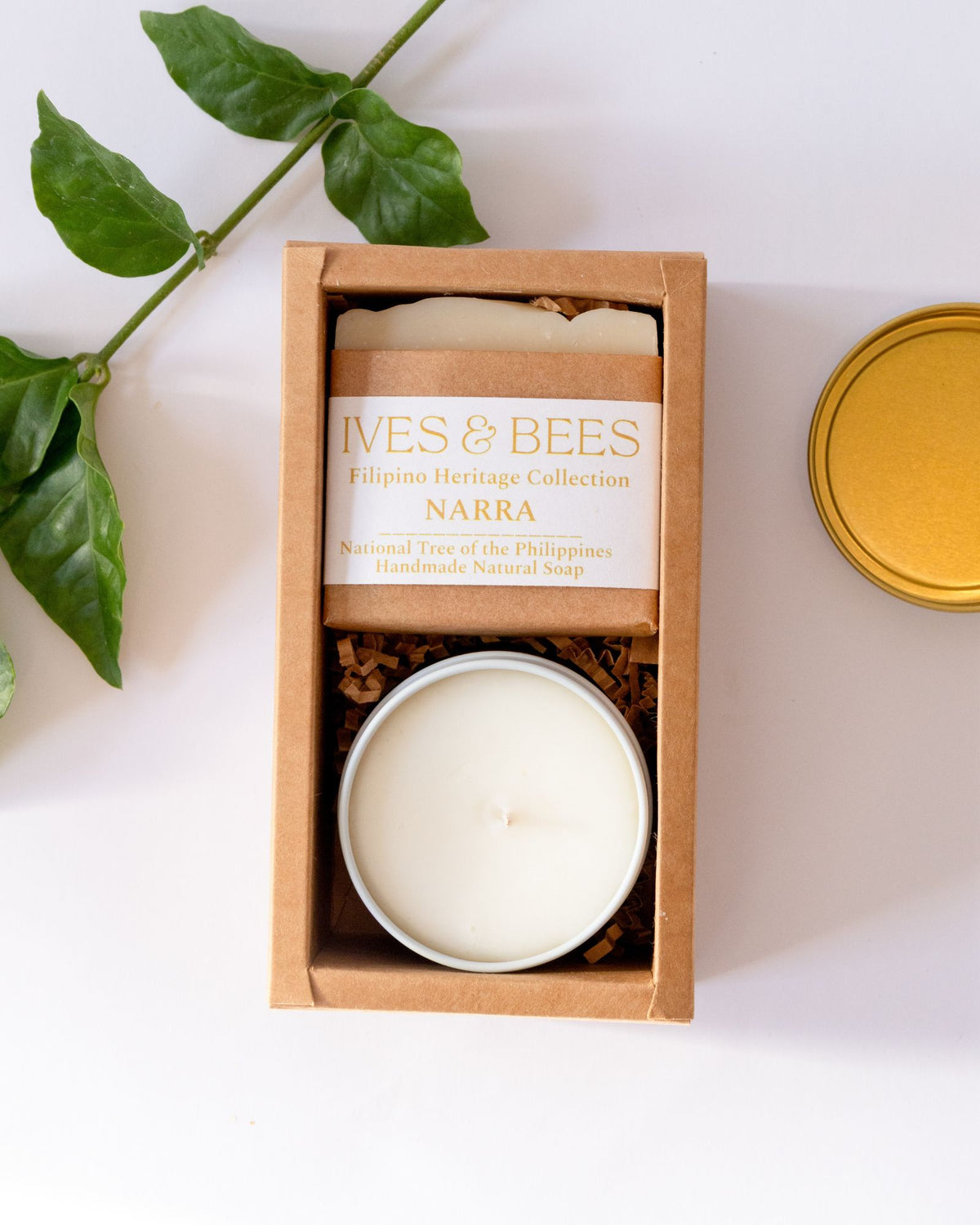 A boxed candle set on a white background with a green leaf sprig and a gold tin lid nearby. The main product is a rectangular kraft paper box containing a round tin candle and a wrapped soap bar.