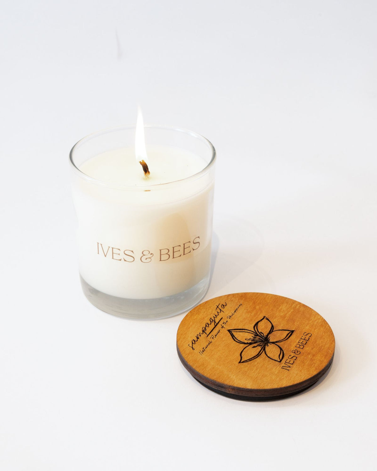 A lit white candle in a clear glass jar with the brand name 'IVES & BEES' printed on the front, shown next to a round wooden lid engraved with a flower and the brand name on a clean white background.