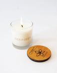 A lit white candle in a clear glass jar with the brand name 'IVES & BEES' printed on the front, shown next to a round wooden lid engraved with a flower and the brand name on a clean white background.