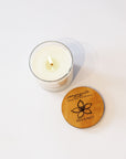 A lit white candle in a clear glass jar sits on a white surface next to its round wooden lid, which is engraved with a flower and the brand name IVES & BEES.