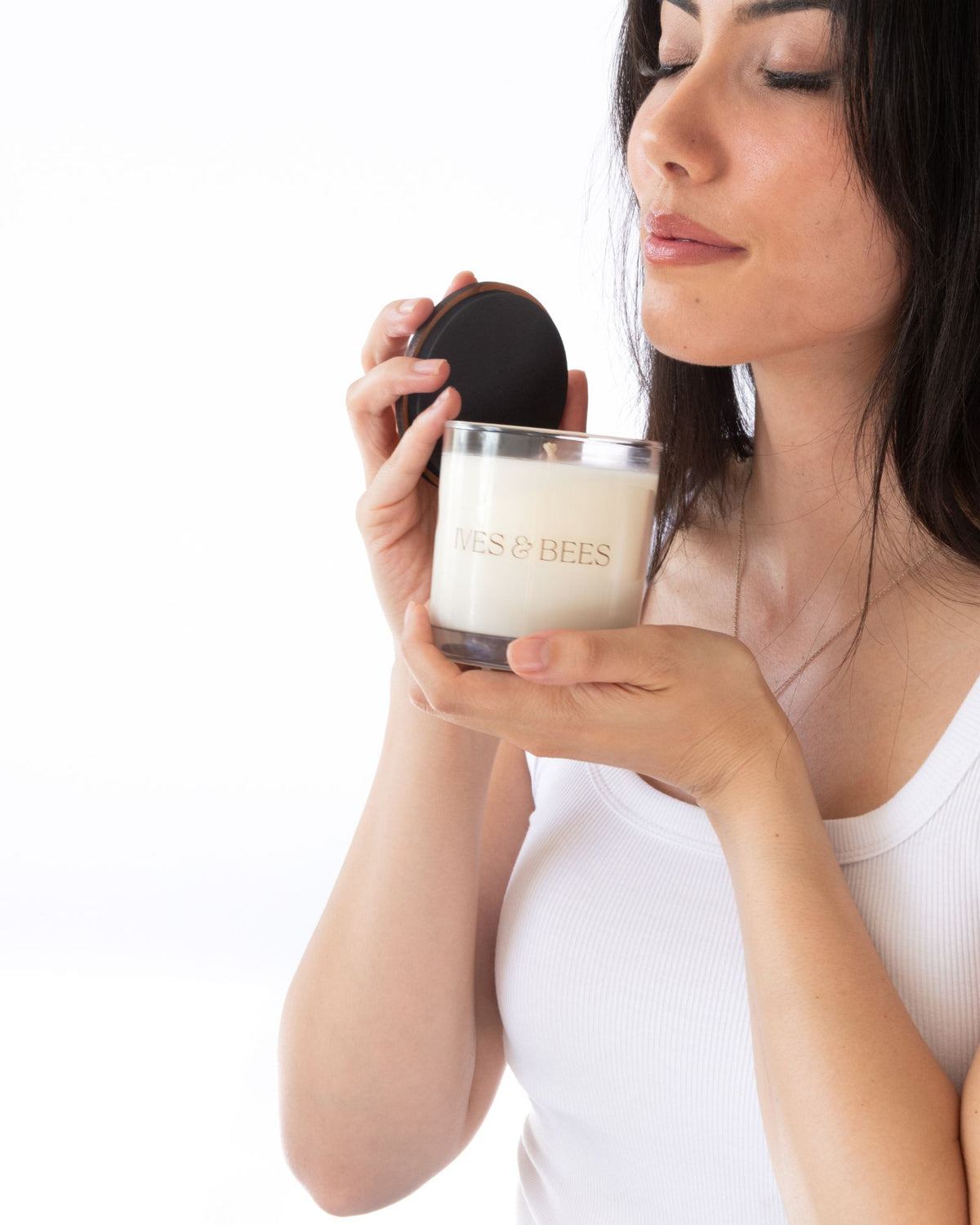 A woman holds a glass jar candle with cream-colored wax and a black lid, with the brand name 'IVES & BEES' printed on the glass, against a solid white background.