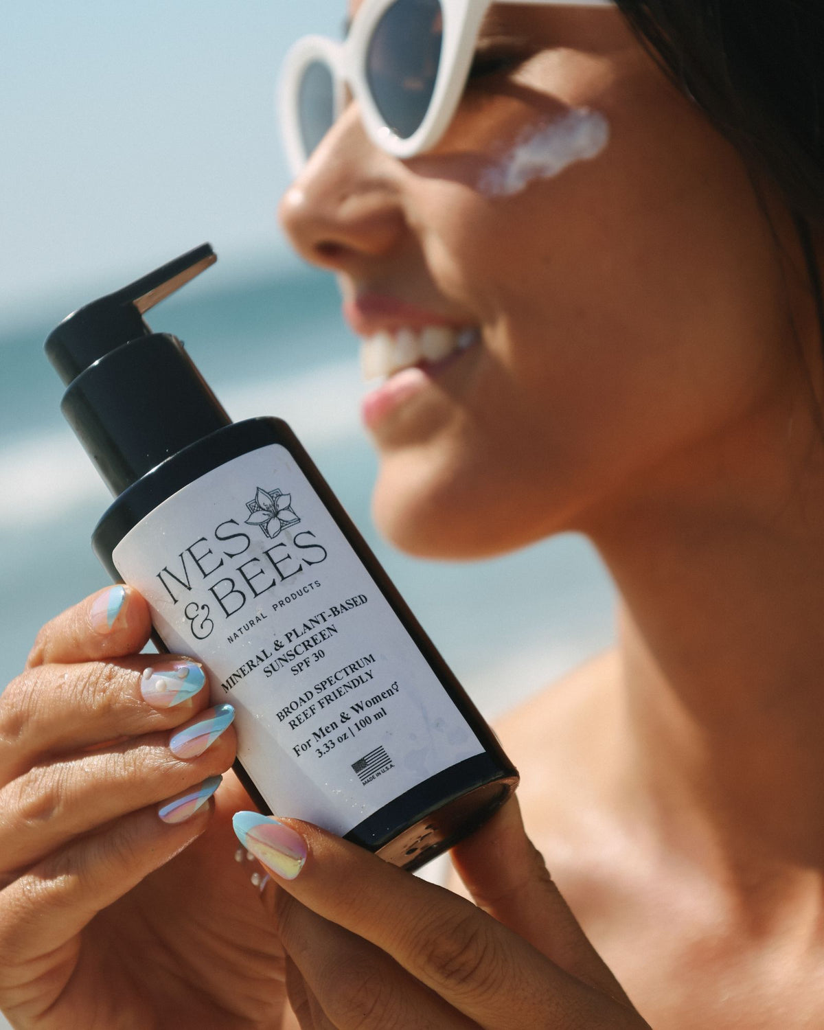 A woman outdoors near the beach holds a bottle of Ives & Bees Sunscreen SPF 50+. She is wearing white sunglasses and has sunscreen on her cheek.