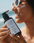 A woman outdoors near the beach holds a bottle of Ives & Bees Sunscreen SPF 50+. She is wearing white sunglasses and has sunscreen on her cheek.
