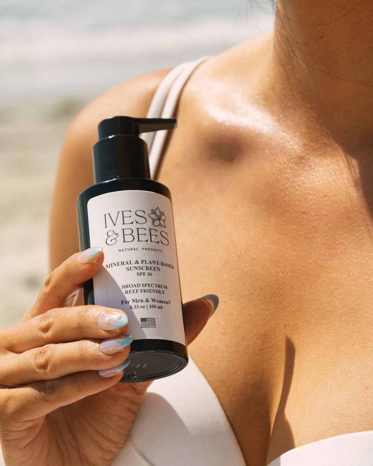 A person outdoors at the beach holds a black pump bottle labeled Ives & Bees Mineral & Plant Rose Sunscreen. 